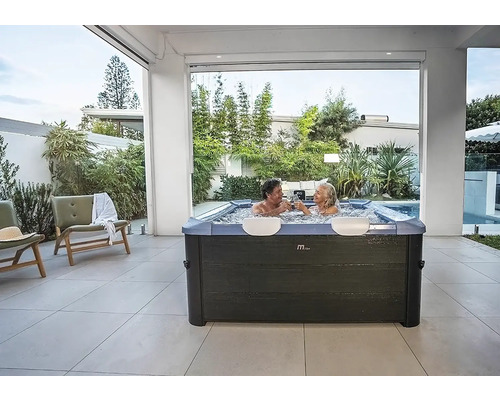Moderner quadratischer Whirlpool mit zwei Personen auf einer hell gefliesten Terrasse inmitten von Gartenpflanzen.