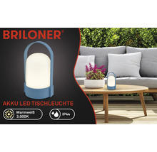 Briloner Logo, Akku-LED-Tischleuchte für außen, warmweißes Licht 3000 Kelvin, IP44 Spritzwasserschutz, tragbare Leuchte mit Henkel auf einem Gartentisch.
