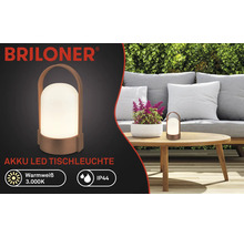Briloner Logo, Akku LED Tischleuchte auf Holztisch in heller Gartenszene mit Sofa und Hecke, warmweiß 3000 Kelvin, spritzwassergeschützt IP44.