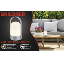 Briloner Logo, Akku LED Tischleuchte, warmweiß 3000 Kelvin, IP44 spritzwassergeschützt, tragbar mit Tragegriff, im Garten auf einem Holztisch.
