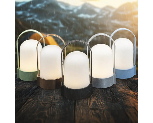 Fünf tragbare Outdoor-LED-Laternen in verschiedenen Farben auf einem Holztisch bei Regen vor einer Gebirgskulisse.