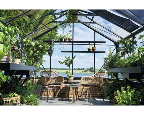 Helles Gewächshaus aus Glas und Metall mit zwei Korbsesseln, einem Holztisch und zahlreichen Pflanzen vor einer Aussicht auf einen See.