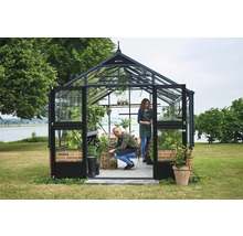 Juliana Gewächshaus aus Glas mit schwarzem Metallrahmen und Ziegelsockel in einem Garten am See. Zwei Personen gärtnern im hellen Innenraum.
