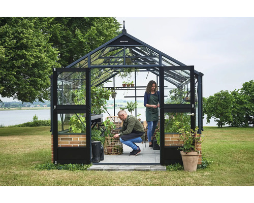 Juliana Gewächshaus aus Glas mit schwarzem Metallrahmen und Ziegelsockel in einem Garten am See. Zwei Personen gärtnern im hellen Innenraum.