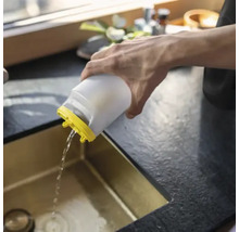 Eine Person gießt Wasser aus einem weißen Kunststoffbehälter mit gelbem Deckel in eine goldfarbene Küchenspüle.