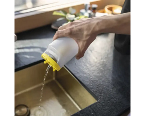 Eine Person gießt Wasser aus einem weißen Kunststoffbehälter mit gelbem Deckel in eine goldfarbene Küchenspüle.