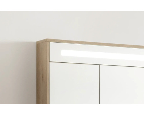 Obere Ecke eines Badezimmerschranks mit Korpus in Eichenoptik, weißer Front und integrierter LED-Beleuchtung.