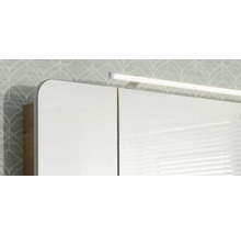 Spiegelschrank mit LED-Aufsatzleuchte und abgerundeten Ecken vor einer gemusterten Wand.