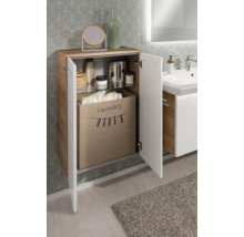 Badezimmerschrank in Holzoptik mit weißen Fronten, geöffneter Tür, Stoff-Wäschesammler und Glasfachboden vor gemusterter Tapete.