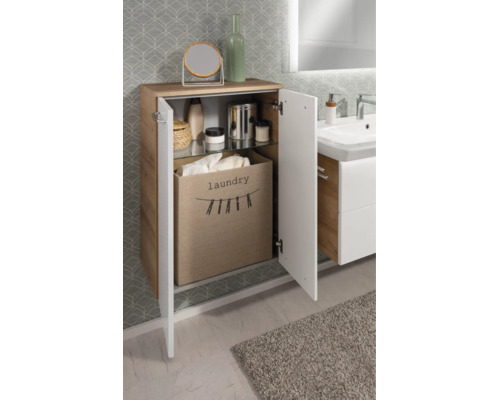 Badezimmerschrank in Holzoptik mit weißen Fronten, geöffneter Tür, Stoff-Wäschesammler und Glasfachboden vor gemusterter Tapete.