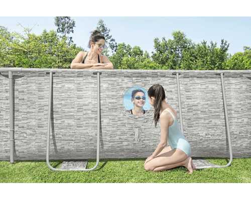 Aufstellpool mit Steinoptik-Außenwand und Sichtfenster im Garten, eine Frau im Pool und ein Mädchen schaut von außen durch das Fenster.