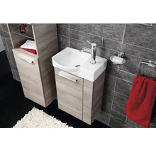 Modernes Badezimmer mit Waschbeckenunterschrank und Hochschrank in Holzoptik, Keramikwaschbecken und dunkler Fliesenwand.
