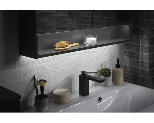 Modernes Badezimmer mit weißem Waschtisch, schwarzer Armatur, beleuchtetem Spiegelschrank und Badaccessoires vor einer strukturierten Wand.