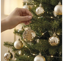 Eine Hand hängt eine goldene Weihnachtskugel an einen grünen Weihnachtsbaum, der mit Lichtern und Christbaumschmuck dekoriert ist.