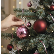 Eine Hand hängt eine glänzende rosa Weihnachtskugel an einen geschmückten Christbaum mit dunkelroten Kugeln und Lichtern.