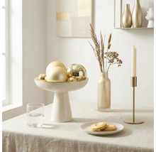 Hell und geordnet gedeckter Tisch mit weißer Schale auf Fuß mit goldenen Kugeln, schmaler Vase mit Trockengräsern und goldenem Kerzenhalter.