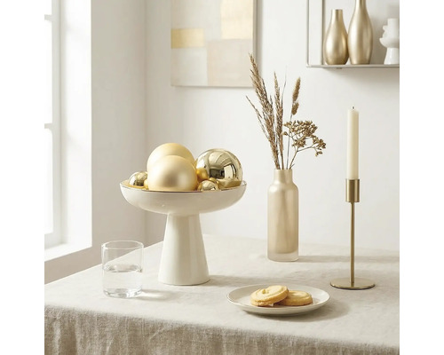 Hell und geordnet gedeckter Tisch mit weißer Schale auf Fuß mit goldenen Kugeln, schmaler Vase mit Trockengräsern und goldenem Kerzenhalter.