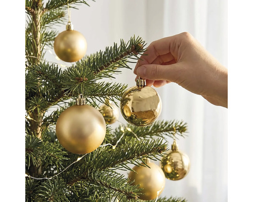 Eine Hand hängt eine glänzende goldene Weihnachtskugel an einen Tannenbaum mit weiteren matten und glänzenden Kugeln.