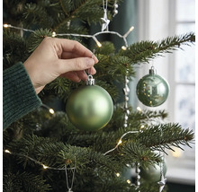 Eine Hand hängt eine matte grüne Weihnachtskugel an einen geschmückten Christbaum mit LED Lichterkette.