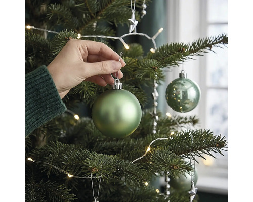 Eine Hand hängt eine matte grüne Weihnachtskugel an einen geschmückten Christbaum mit LED Lichterkette.
