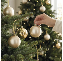 Eine Hand platziert eine champagnerfarbene Weihnachtskugel an einem hell erleuchteten, grünen Christbaum in festlicher Atmosphäre.