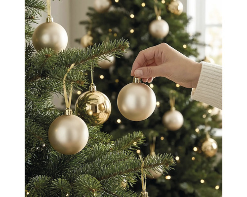 Eine Hand platziert eine champagnerfarbene Weihnachtskugel an einem hell erleuchteten, grünen Christbaum in festlicher Atmosphäre.