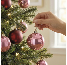 Eine Hand hängt eine rosa glänzende Weihnachtskugel an einen Weihnachtsbaum mit warmweißen Lichtern und bordeauxrotem Baumschmuck.