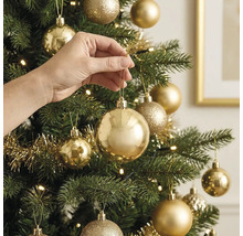 Hand hängt eine glänzende goldene Weihnachtskugel an einen künstlichen Weihnachtsbaum mit weiterem Goldschmuck und Lichtern.