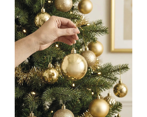 Hand hängt eine glänzende goldene Weihnachtskugel an einen künstlichen Weihnachtsbaum mit weiterem Goldschmuck und Lichtern.