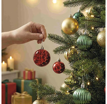 Eine Hand hängt eine rote Weihnachtskugel an einen geschmückten Christbaum mit Lichtern und Geschenken im Hintergrund.