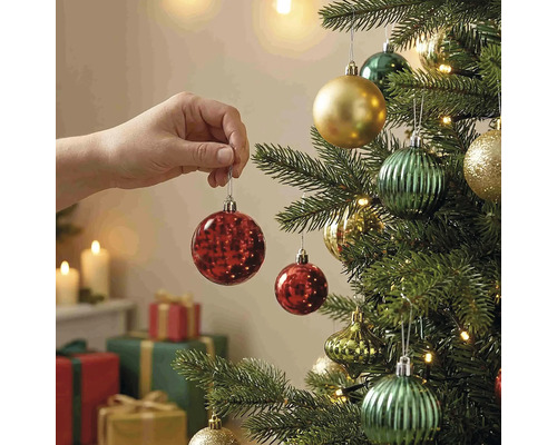 Eine Hand hängt eine rote Weihnachtskugel an einen geschmückten Christbaum mit Lichtern und Geschenken im Hintergrund.
