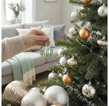Eine Hand hängt eine gerippte grüne Weihnachtskugel aus Glas an einen geschmückten Christbaum in einem hellen Wohnzimmer.
