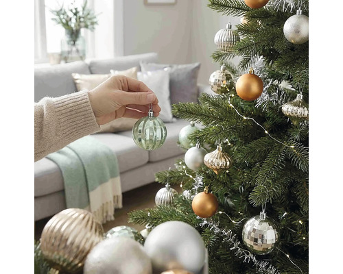 Eine Hand hängt eine gerippte grüne Weihnachtskugel aus Glas an einen geschmückten Christbaum in einem hellen Wohnzimmer.