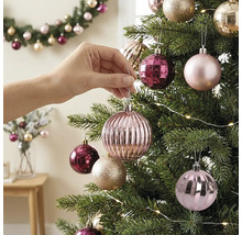 Hand hängt eine rosa geriffelte Weihnachtskugel an einen geschmückten Tannenbaum mit Lichterkette.