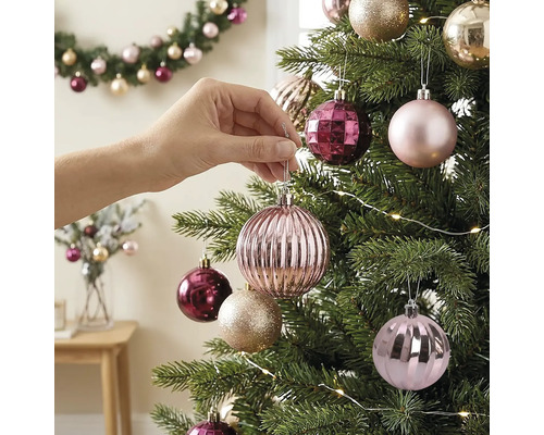 Hand hängt eine rosa geriffelte Weihnachtskugel an einen geschmückten Tannenbaum mit Lichterkette.
