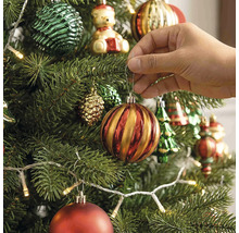 Eine Hand hängt eine gestreifte Weihnachtskugel an einen geschmückten Weihnachtsbaum mit Lichterkette und verschiedenen Ornamenten.