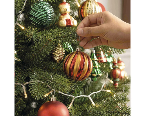 Eine Hand hängt eine gestreifte Weihnachtskugel an einen geschmückten Weihnachtsbaum mit Lichterkette und verschiedenen Ornamenten.