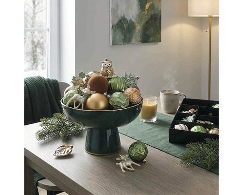 Festliche Tischdekoration mit einer dunkelgrünen Fußschale voller Weihnachtsanhänger, einer Kerze und Tannenzweigen auf einem Holztisch.