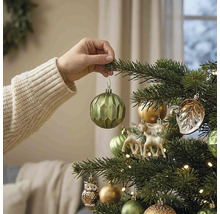 Hand hängt eine grüne geometrische Weihnachtskugel an einen geschmückten Weihnachtsbaum mit Hirsch- und Eulenfiguren sowie warmen Lichtern.