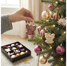 Eine Hand hängt einen engelsförmigen Anhänger an einen beleuchteten Weihnachtsbaum mit Kugeln und Sternen neben einer Box mit weiterem Baumschmuck.