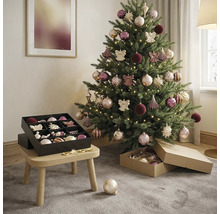 Geschmückter Weihnachtsbaum in hellem Zimmer mit warmweißem Licht, Kugeln in Rosa und Lila sowie schwarzer Aufbewahrungsbox auf einem Holzhocker.
