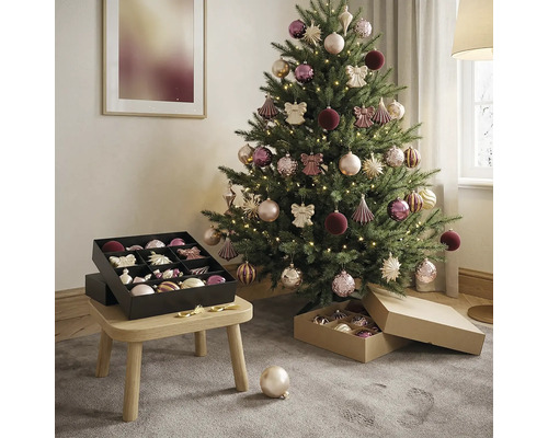 Geschmückter Weihnachtsbaum in hellem Zimmer mit warmweißem Licht, Kugeln in Rosa und Lila sowie schwarzer Aufbewahrungsbox auf einem Holzhocker.