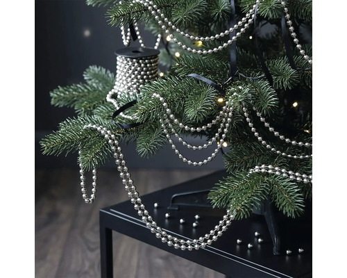 Silberne Perlenkette an den Zweigen eines Christbaums mit warmweißen Lichtern.
