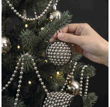 Eine Hand hängt eine runde Christbaumkugel aus vielen kleinen silbernen Perlen an einen geschmückten Weihnachtsbaum mit Lichterkette.