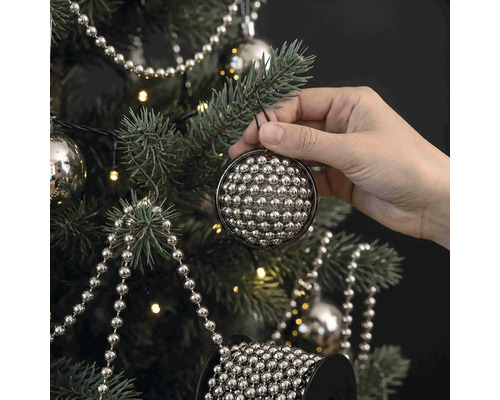 Eine Hand hängt eine runde Christbaumkugel aus vielen kleinen silbernen Perlen an einen geschmückten Weihnachtsbaum mit Lichterkette.