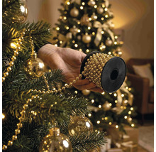 Hand hält Spule mit Perlenkette beim Schmücken eines Weihnachtsbaums mit Christbaumkugeln und Lichtern.