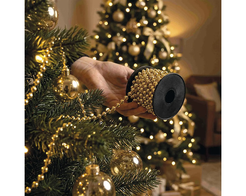 Hand hält Spule mit Perlenkette beim Schmücken eines Weihnachtsbaums mit Christbaumkugeln und Lichtern.