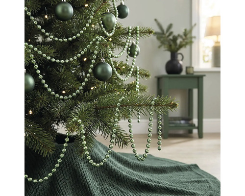 Dekorierter Weihnachtsbaum mit matten Kugeln, einer Perlenkette und einer gestrickten Weihnachtsbaumdecke in einem Wohnzimmer.