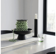 Grüne Perlenkette auf einer schwarzen Dekoschale, neben einem schwarzen Kerzenhalter und einem Wasserglas auf einem hellen Tisch.