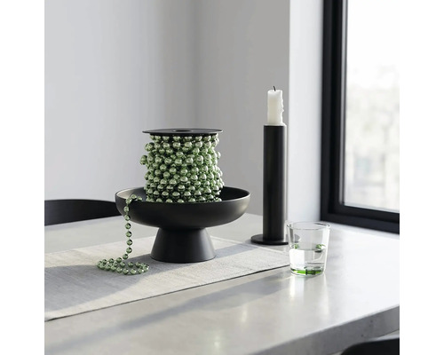Grüne Perlenkette auf einer schwarzen Dekoschale, neben einem schwarzen Kerzenhalter und einem Wasserglas auf einem hellen Tisch.